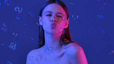 Bir kadın neşeli bir ifadeyle poz verir, dudaklarını büzer ve göz kırpar, mavi bir arkaplanda durur..