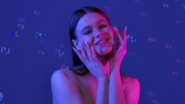 Bir kadın, mavi bir arka planda etrafında yüzen sabun köpükleri gibi neşeyle gülümsüyor, bir neşe ve kapris hissi uyandırıyor. Yavaş çekim