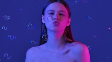 Bir kadın neşeli bir ifadeyle poz verir, dudaklarını büzer ve göz kırpar, mavi bir arka planda durur. Yavaş çekim