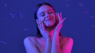 Bir kadın, mavi bir arka planda etrafında yüzen sabun köpükleri gibi neşeyle gülümsüyor, bir neşe ve kapris hissi uyandırıyor. Yavaş çekim