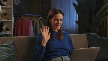 Mavi kazaklı mutlu bir kadın dizüstü bilgisayarıyla konuşurken gülümsüyor, koltukta rahatça oturuyor..