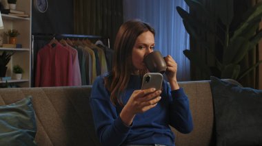Kanepede oturan, kahve içen, rahat bir oturma odasında akıllı telefonuyla video izleyen bir kadın..