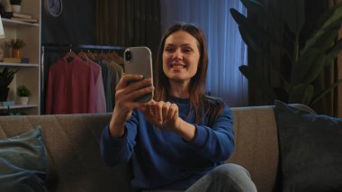 Mavi kazaklı mutlu bir kadın telefonuyla konuşurken gülümsüyor, koltukta rahatça oturuyor..