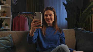 Mavi kazaklı mutlu bir kadın telefonuyla konuşurken gülümsüyor, koltukta rahatça oturuyor..