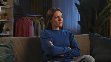 Mavi kazaklı bir kadın kanepede oturuyor, kolları bağlı, dalgın görünüyor. Elbise askısı ve bitkiler görünür..