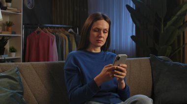 Kanepede oturan ve rahat bir oturma odasında akıllı telefonuyla video izleyen bir kadın..