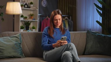 Kanepede oturan ve rahat bir oturma odasında akıllı telefonuyla video izleyen bir kadın..