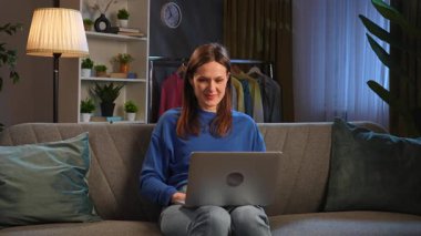 Mavi kazaklı mutlu bir kadın dizüstü bilgisayarıyla konuşurken gülümsüyor, koltukta rahatça oturuyor..