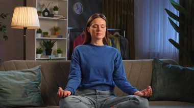 Huzurlu bir oturma odasındaki kanepede meditasyon yapan bir kadın. Etrafı bitkilerle çevrili ve dinlenmek için hafif ışıklandırmalı..