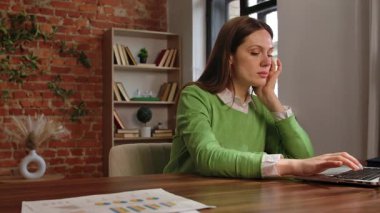 Yeşil kazaklı bir kadın, ev ofisinin duvarları tuğladan yapılmış bir bilgisayarında çalışırken stresli görünür..