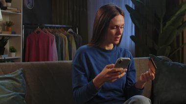 Kanepede oturan ve rahat bir oturma odasında akıllı telefonuyla video izleyen bir kadın..