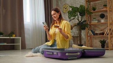 Evde yerde oturan bir kadın, akıllı telefon kullanıyor ve bir bavulun yanında kredi kartı tutuyor..