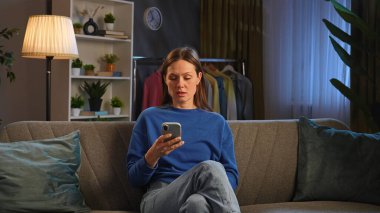 Kanepede oturan ve rahat bir oturma odasında akıllı telefonuyla video izleyen bir kadın..