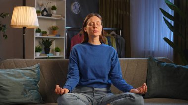 Huzurlu bir oturma odasındaki kanepede meditasyon yapan bir kadın. Etrafı bitkilerle çevrili ve dinlenmek için hafif ışıklandırmalı..