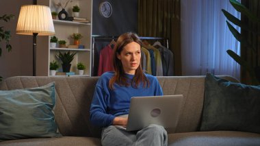 Bir kadın rahat bir odada dizüstü bilgisayarla kanepede oturuyor. Bitkiler, giysiler ve yumuşak ışıklandırma sıcak bir atmosfer yaratır..