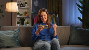Bir kadın, akıllı telefonuyla eğlendirici bir şey izlerken gülümseyerek kanepede öylesine oturur..