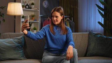 Mavi kazaklı mutlu kadın koltukta rahatça otururken gülümsüyor ve cep telefonuyla selfie çekiyor..