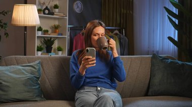 Bir kadın rahat bir şekilde kanepede oturur, akıllı telefonuyla uğraşırken kahvesini yudumlar, rahatlamanın tadını çıkarır..
