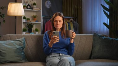 Bir kadın rahat bir şekilde kanepede oturur, akıllı telefonuyla uğraşırken kahvesini yudumlar, rahatlamanın tadını çıkarır..