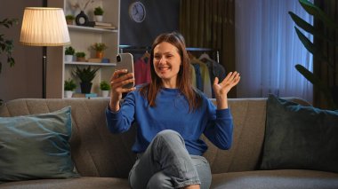 Mavi kazaklı mutlu bir kadın telefonuyla konuşurken gülümsüyor, koltukta rahatça oturuyor..