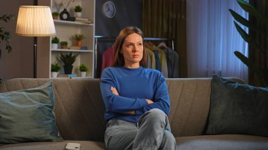 Mavi kazaklı bir kadın kanepede oturuyor, kolları bağlı, dalgın görünüyor. Elbise askısı ve bitkiler görünür..