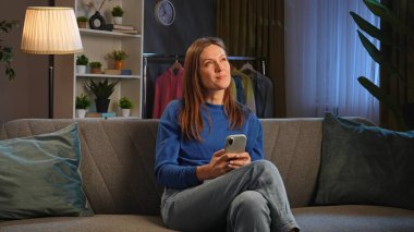 Kanepede oturan ve rahat bir oturma odasında akıllı telefonuyla video izleyen bir kadın..
