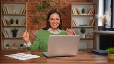 Yeşil kazaklı kadın, ev bilgisayarında çalışırken mutlu görünüyor. Duvarları tuğladan..