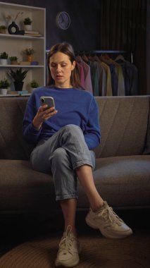 Kanepede oturan ve rahat bir oturma odasında akıllı telefonuyla video izleyen bir kadın..