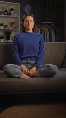 Huzurlu bir oturma odasındaki kanepede meditasyon yapan bir kadın. Etrafı bitkilerle çevrili ve dinlenmek için hafif ışıklandırmalı..