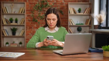 Yeşil kazaklı bir kadın telefonuna bakar ve hesap makinesi ve dizüstü bilgisayar kullanarak masasında çalışır..