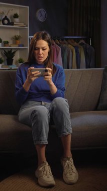 Bir kadın rahat bir şekilde kanepede oturuyor, üzgün ve akıllı telefonundan bir şeye bakıyor..