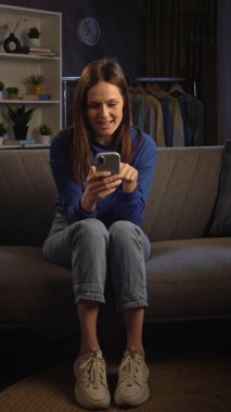 Bir kadın rahat bir şekilde kanepede oturuyor, üzgün ve akıllı telefonundan bir şeye bakıyor..