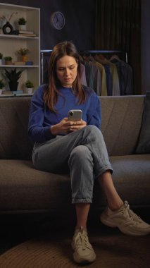 Kanepede oturan ve rahat bir oturma odasında akıllı telefonuyla video izleyen bir kadın..