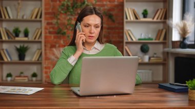 Yeşil kazaklı bir kadın telefonla konuşuyor ve masasında hesap makinesi ve dizüstü bilgisayar kullanarak çalışıyor. Tuğla duvara karşı duran belgelerin arasında..
