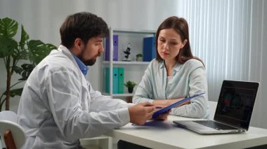Tıp bürosunda bir hastaya danışmanlık yapan bir doktor. Masada dizüstü bilgisayar ve şişe gibi çeşitli eşyalar var..
