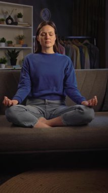 Huzurlu bir oturma odasındaki kanepede meditasyon yapan bir kadın. Etrafı bitkilerle çevrili ve rahatlamak için hafif ışıklandırılmış. Dikey video