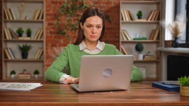 Yeşil kazaklı bir kadın, tuğla duvarlı bir ofiste dizüstü bilgisayarla çalışırken düşünceli görünür..