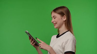 Gençlik duyguları konsepti. Kameranın önünde poz veren, gülümseyen, tablet tutan ve mesaj yazan genç bir kız. Renkli yeşil ekran stüdyo arka plan modelinde izole edilmiş. Yavaş çekim