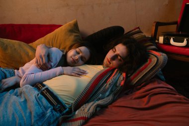 İki insan rahat bir yatakta rahatlar. Etrafları minderlerle çevrili ve sıcak, davetkar bir atmosfer yaratıyorlar..