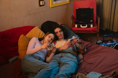 İki arkadaş rahat bir yatakta oturmuş video oyunları oynuyorlar. Sıcak atmosfer yumuşak ışıklandırma ve renkli sanat eserlerini içerir..