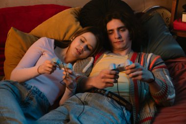 İki arkadaş rahat bir yatakta oturmuş video oyunları oynuyorlar. Sıcak atmosfer yumuşak ışıklandırma ve renkli sanat eserlerini içerir..