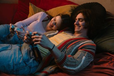 İki arkadaş rahat bir yatakta oturmuş video oyunları oynuyorlar. Sıcak atmosfer yumuşak ışıklandırma ve renkli sanat eserlerini içerir..