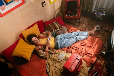 Genç bir adam rahat bir şekilde uzanıyor, rahat bir odada klasik bir oyun konsolu ile meşgul. Üst çekim