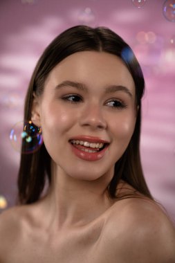 Orta boylu, kahverengi saçlı genç bir kadın neşeli bir şekilde gülümser. Etrafı kabarcıklarla ve pembe bir arka planla çevrilidir..