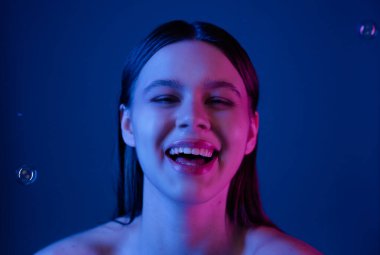 Bir kadın neşeyle gülüyor, koyu bir arkaplanda pembe ve mavi ışıklarla aydınlanıyor..