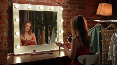 Kırmızı elbiseli bir kadın şık bir soyunma odasında, yanan aynanın önünde makyaj yapar..