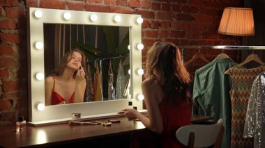 Kırmızı elbiseli bir kadın, şık ışıklandırması ve şık kıyafetleri olan bir soyunma odasına makyaj yapar..