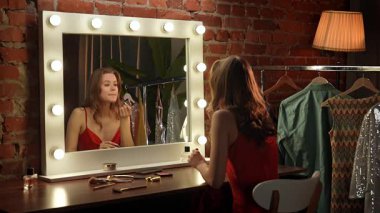 Bir kadın parlak ışıklarla çevrili büyük bir aynanın önünde ruj sürerken görülüyor..