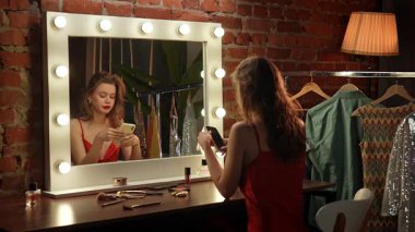 Kırmızı ruj ve altın küpeli genç bir kadın akıllı telefonuna bakarken gülümsüyor. Arkasında, askıdaki birkaç elbise ve bazı yeşil bitkiler görülüyor..