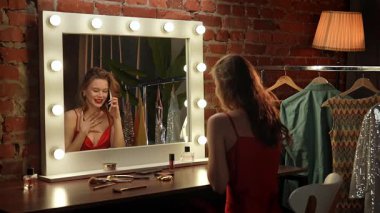 Kırmızı elbiseli bir kadın şık kıyafetlerle çevrili gösterişli bir aynanın önünde makyaj yapar..